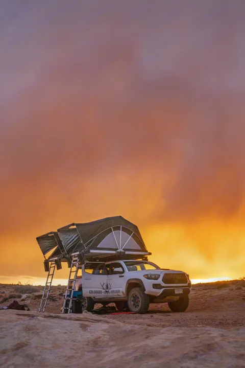 RV rental adventure: White Toyota truck with rooftop tent set up at a vibrant sunset campsite on rocky terrain.. Toyota Tacoma 2022
