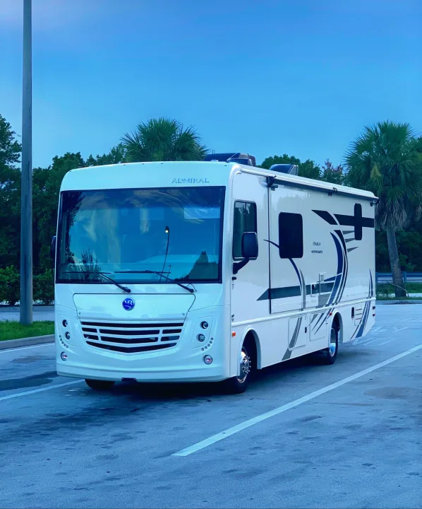 A modern white Admiral RV rental parked outdoors. Clear blue sky, green trees & palm trees visible in background.. Holiday Rambler Admiral 2021