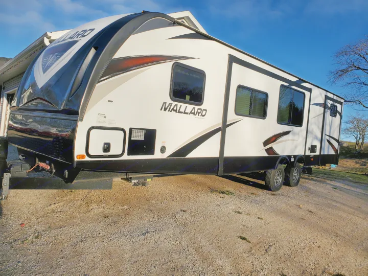White Heartland Mallard travel trailer, ideal for an RV rental vacation. Parked on gravel under a clear blue sky.. Heartland RVs Mallard 2019