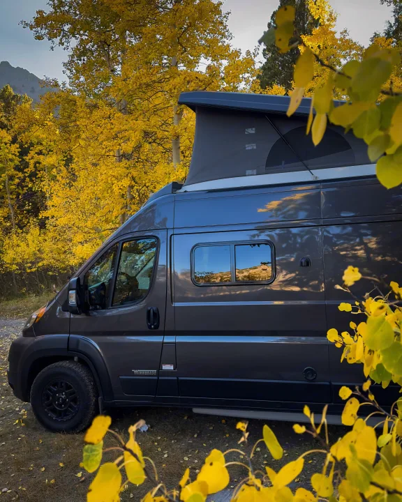 Grey RV rental with pop-top tent immersed in vibrant golden aspen trees, reflecting mountains & sky, perfect for fall adventures.. Winnebago Solis 59PX 2023