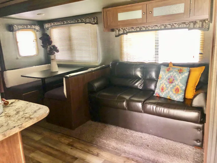 RV rental interior showing a dinette, dark leather couch, overhead cabinets, and sunlit windows.. Keystone RV Hideout 2019