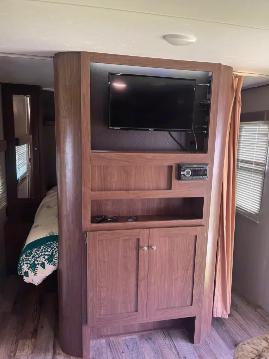 RV rental interior showing a compact entertainment center with TV, stereo, storage, and a glimpse into the connected bedroom area.. Keystone RV Hideout 2019