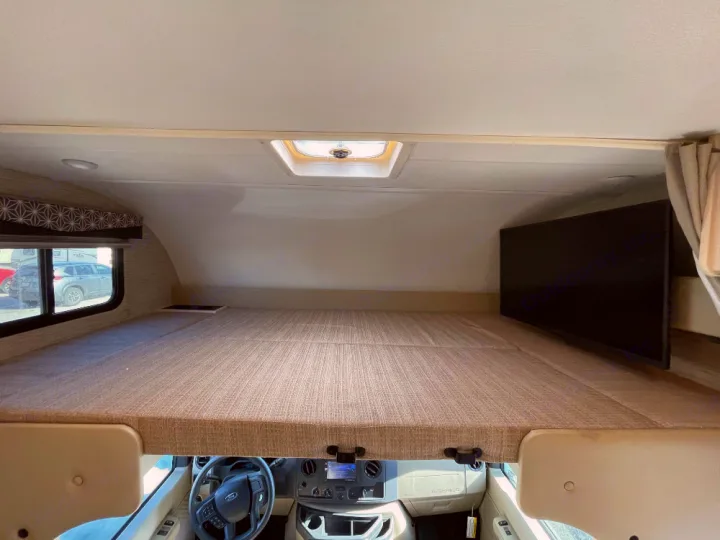 Inside an RV rental: a comfortable cab-over bunk bed featuring a TV, window, and skylight, with the cockpit below.. Thor Freedom Elite 2022