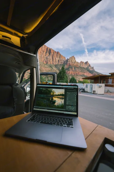 RV rental's mobile office with a laptop editing photos, framed by spectacular red rock mountains. A digital nomad's workspace.. Ram Promaster 2021