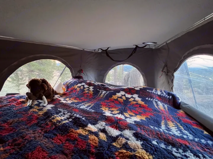 Cozy RV rental pop-top tent bed with colorful blanket, plush toys, and arched windows revealing a forest view.. Winnebago Solis 2022