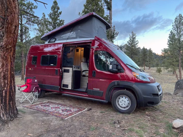 Red Winnebago Solis campervan with pop-top extended & side open, a rug & chair outside in a forest setting. Great rv rental!. Winnebago Solis 2022