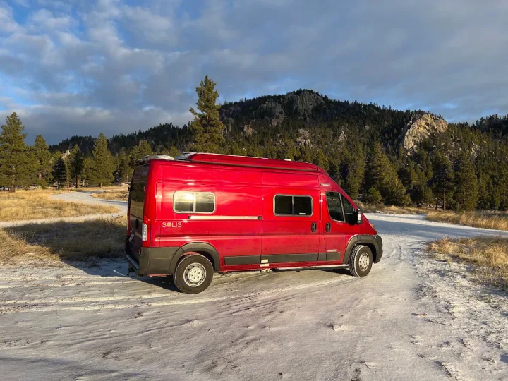 Bright red Winnebago Solis rv rental van on a snowy mountain road, surrounded by pine trees under a cloudy sky.. Winnebago Solis 2022