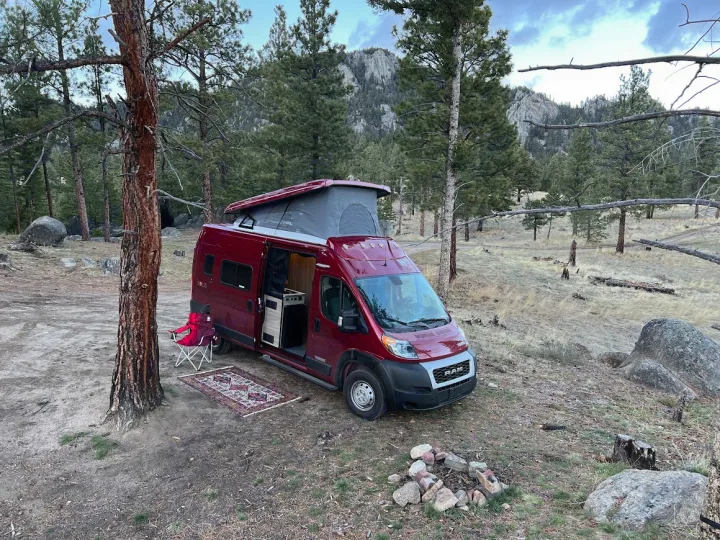 Red pop-top camper van, available for RV rental, set up at a scenic mountain forest campsite with a cozy rug.. Winnebago Solis 2022