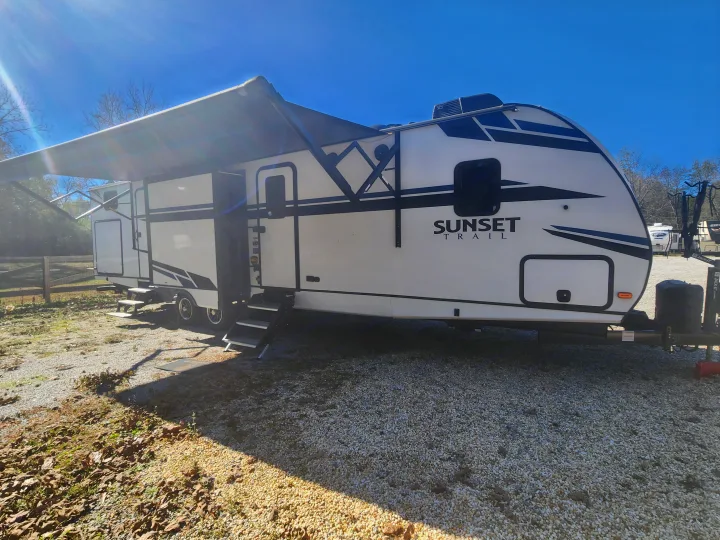 A white and black Sunset Trail RV rental is shown parked on gravel with its awning extended and steps down.. Crossroads Sunset Trail 2021