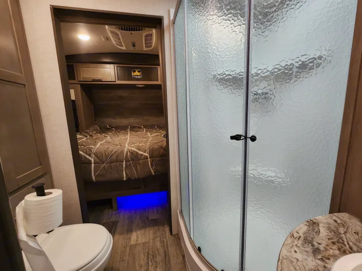 This RV rental features a compact bathroom with toilet and frosted shower, next to a bedroom with a "KING BED" and blue underglow.. Crossroads Sunset Trail 2021