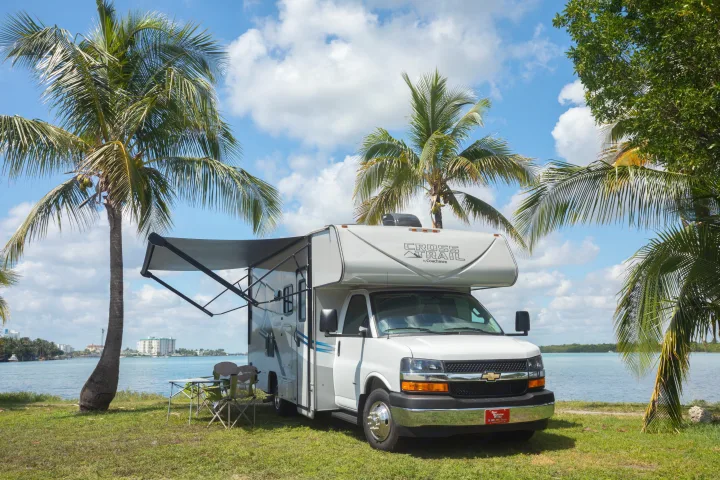Serene waterfront RV rental. A white motorhome with extended awning, chairs, and palm trees by a bay under a blue sky.. Forest River Coachmen Cross Trail XL 2022
