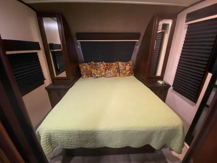 RV rental bedroom featuring a queen bed with light green bedding, patterned pillows, built-in storage, and dark blinds.. Jayco White Hawk 2019