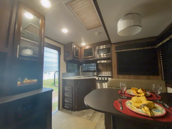 RV rental interior: A full kitchen with dark cabinets, stainless appliances, and a set dining area with tableware under bright lights.. Jayco White Hawk 2019