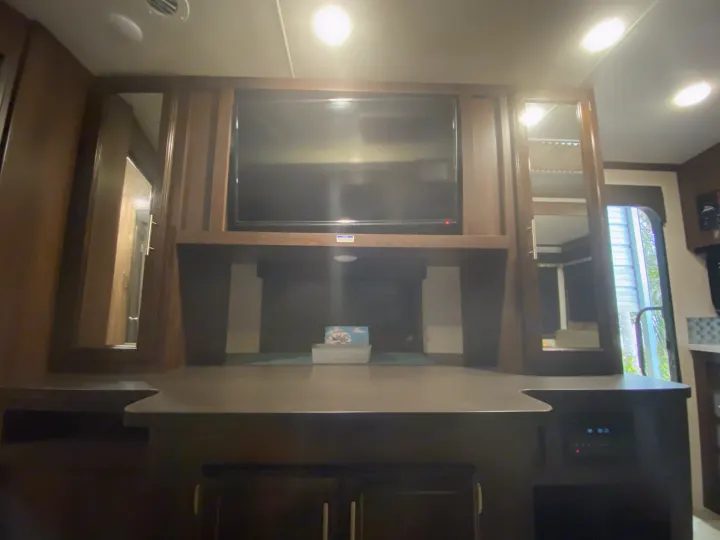 Inside an RV rental: a TV mounted above a counter with controls, flanked by mirrored cabinets reflecting the interior.. Jayco White Hawk 2019