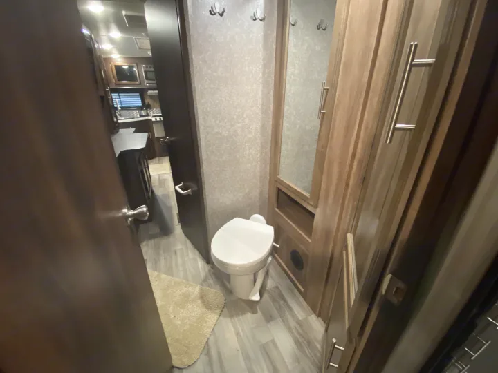 RV rental interior: A compact bathroom with a toilet, mirror, and hooks, leading to the kitchen and living area.. Jayco White Hawk 2019