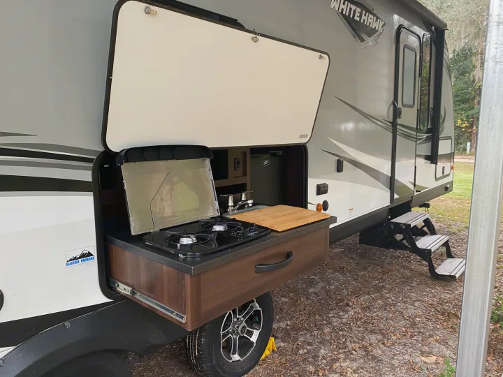 RV rental with a side panel open, revealing a slide-out outdoor kitchen featuring a stove, sink, and mini-fridge.. Jayco White Hawk 2019