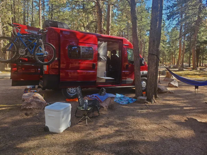 Red RV rental van with bikes parked in a sunlit pine forest campsite. A blue hammock hangs nearby, with a small dog and cooler visible.. Winnebago Solis Pocket 2022
