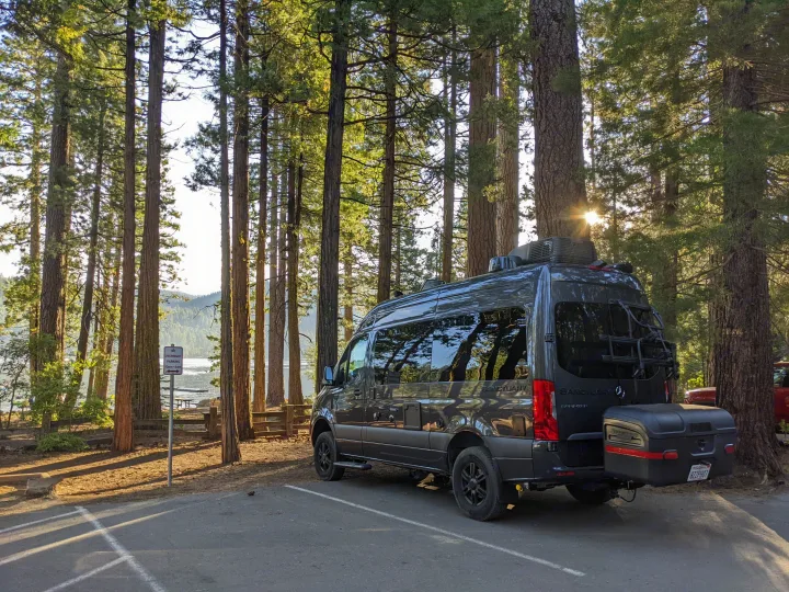 A gray rv rental camper van sits parked by a sunny lake and tall forest trees, hinting at outdoor adventures.. Thor Sanctuary 2022