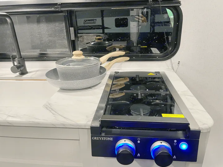 Inside an RV rental, a sleek compact kitchen features a modern stovetop with glowing blue knobs, sink, and nested cookware on white counters.. Forest River Salem FSX 2021