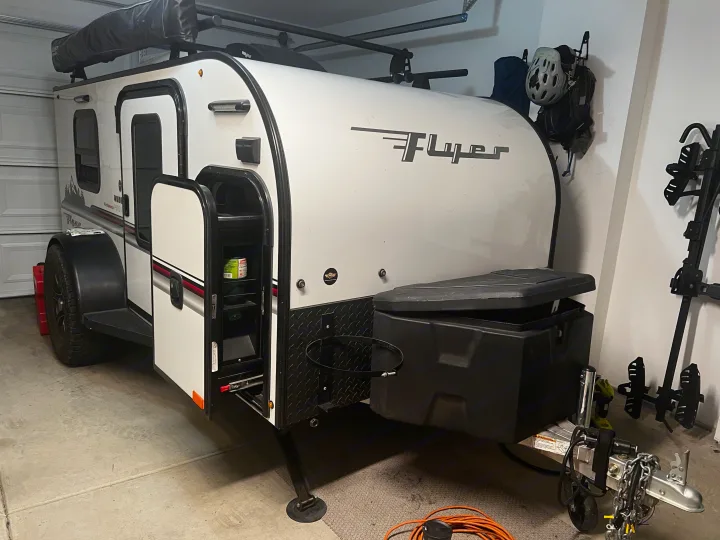 A white Flyer teardrop camper, a compact rv rental, parked in a garage. It features roof racks and an open side utility door.. Custom Camper 2021