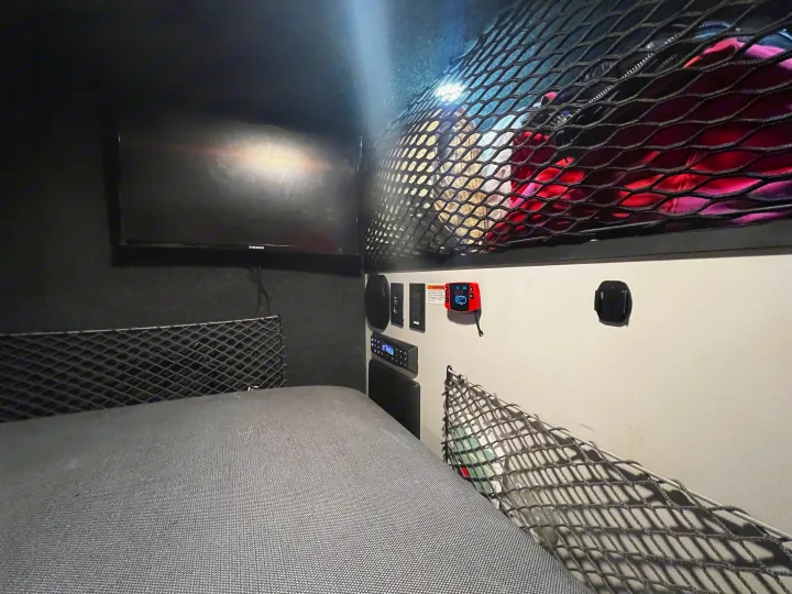 Inside an RV rental: a compact bedroom with a bed, TV, and storage nets overhead and along walls.. Custom Camper 2021