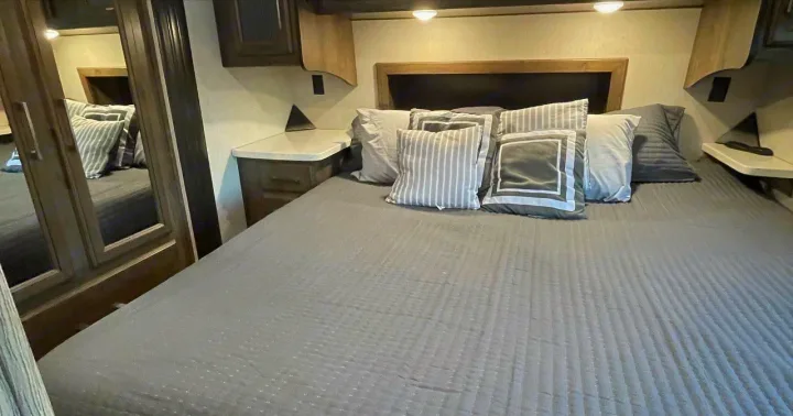 Cozy RV rental bedroom featuring a spacious grey bed, decorative pillows, and built-in nightstands. Mirrored wardrobe on left.. Heartland Cyclone 2018