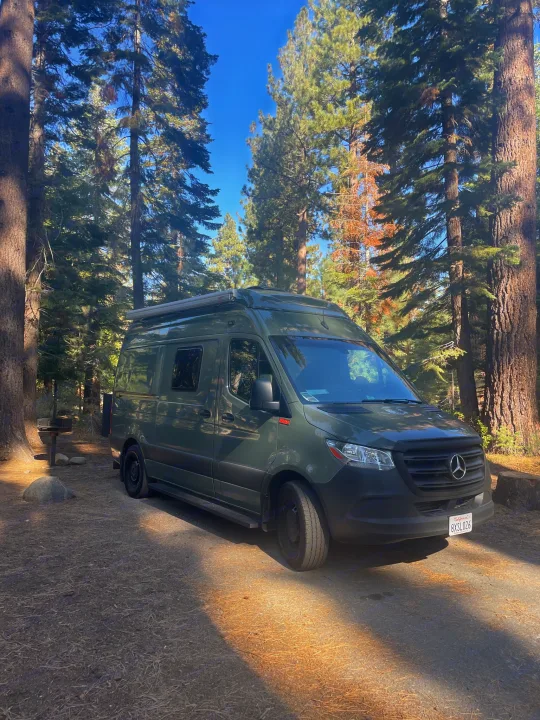 A dark green Sprinter RV rental is parked on a dirt path in a sunny forest campground among tall trees.. Mercedes-Benz Sprinter “144” 2020