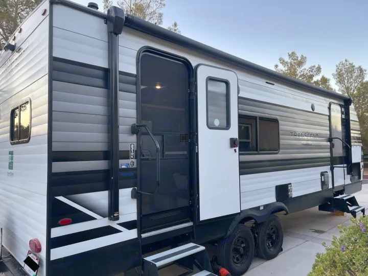 This modern white, black, and gray travel trailer with its door open and steps extended, is ready for your next rv rental adventure.. Grand Design Transcend Xplor 2022