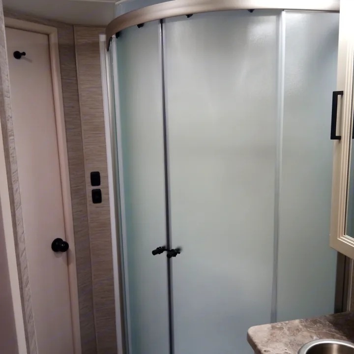 Inside an RV rental, a modern bathroom with a curved frosted glass shower, white door, and vanity with sink.. Thor Four Winds 2022