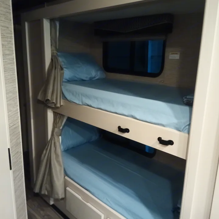 Inside an RV rental: cozy bunk beds with light blue sheets, windows, and curtains for comfortable sleeping.. Thor Four Winds 2022