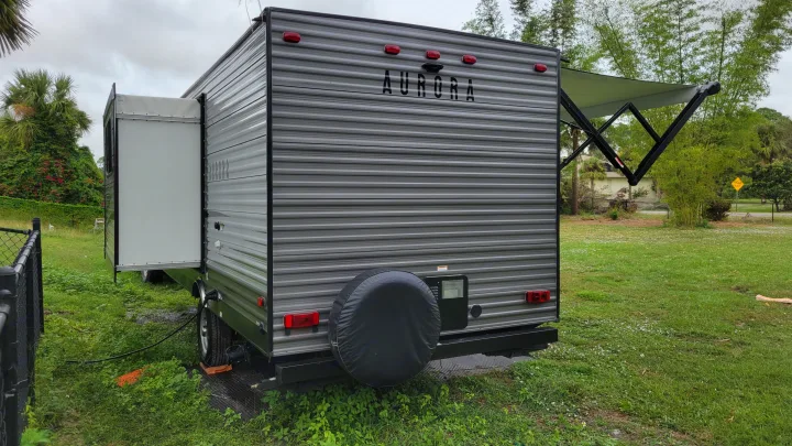 This gray Aurora travel trailer, ready for an RV rental adventure, shows its rear, pop-out, and extended awning in a grassy spot.. Forest River Coachmen Aurora 2022