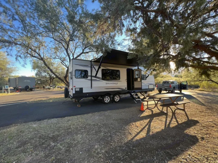 An RV rental travel trailer is parked at a sunny campsite, complete with a truck, picnic table, and shade trees.. Keystone RV Springdale 2020