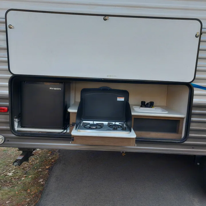 Open compartment of an RV rental revealing a compact outdoor kitchen with a sink, two-burner stove, and fridge.. Avenger Avenger Trailer 2021