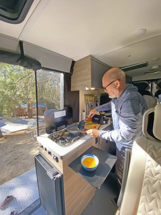 Man cooks eggs in his RV rental kitchen, with a bowl of beaten eggs nearby. A sunny campsite and trees are visible outside the window.. Winnebago Solis 2023