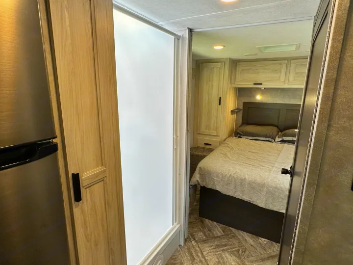 This RV rental interior showcases a cozy bedroom with overhead storage, a sleek shower door, and a modern refrigerator.. Coachmen Sunseeker 2022