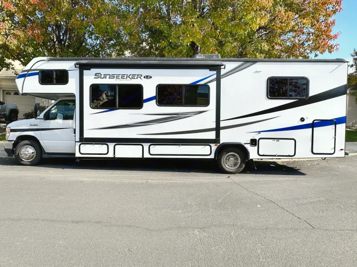 White Class C Sunseeker RV available for rental, featuring blue and black graphics. Parked on a sunny street.. Coachmen Sunseeker 2022