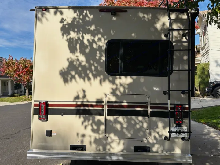 Rear of an RV rental: beige exterior, black ladder, window, California license plate, taillights & backup camera. Ready for travel!. Coachmen Leprechaun 2022