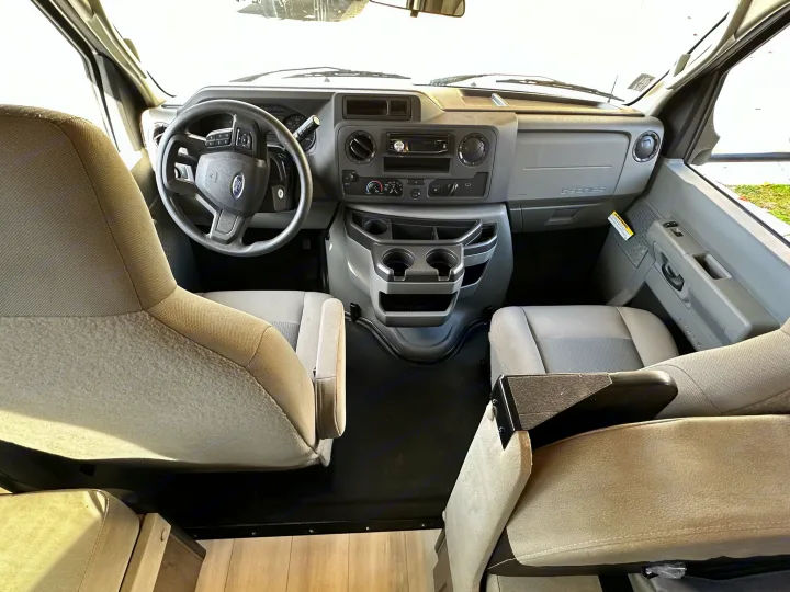Inside an RV rental: a modern, clean cockpit with driver's area, passenger seat, dashboard, and steering wheel.. Coachmen Leprechaun 2022