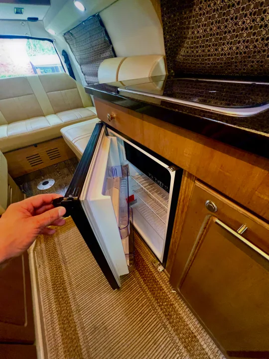 Inside an RV rental, a hand opens a compact refrigerator. The spacious interior features light seating, wood cabinetry, and patterned details.. Roadtrek Roadtrek Class B 2015