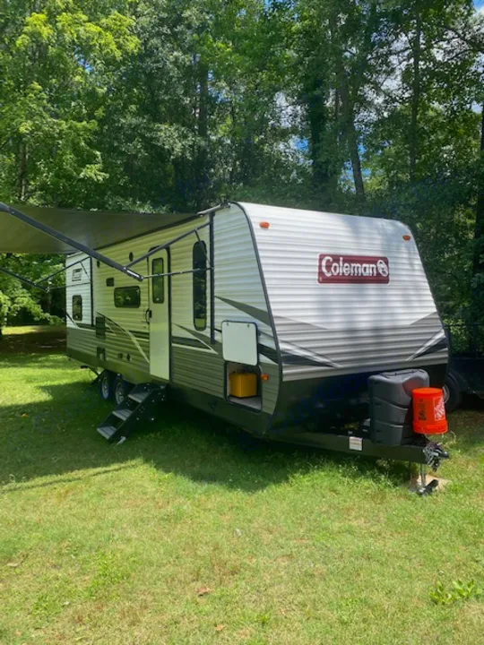 Coleman RV rental ready for adventure. This travel trailer is parked on grass surrounded by trees with its awning extended.. Keystone RV CMMM262BH21 2021