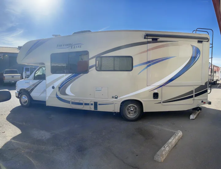 Side view of a beige Freedom Elite Class C RV rental with blue graphics, parked outdoors on a sunny day. Ready for your adventure.. Thor Motor Coach Freedom Elite 2019
