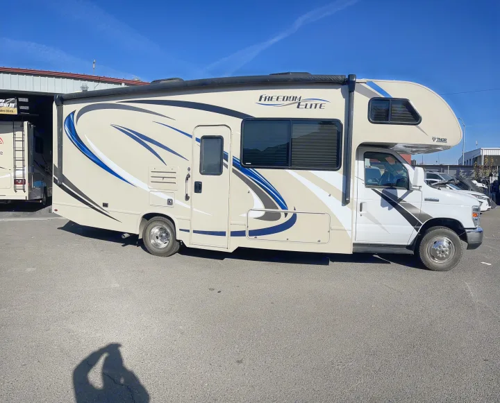 A Class C Thor Freedom Elite RV, ideal for rv rental, parked on asphalt. Beige & white with blue graphics under clear sky.. Thor Motor Coach Freedom Elite 2019