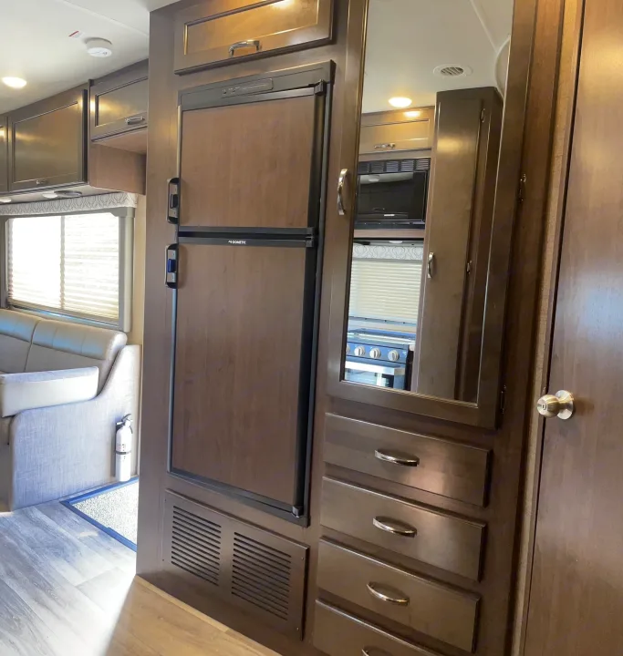 RV rental interior: a functional kitchenette with dark wood fridge, cabinets, and drawers. Light couch by window, fire extinguisher visible.. Thor Motor Coach Freedom Elite 2019