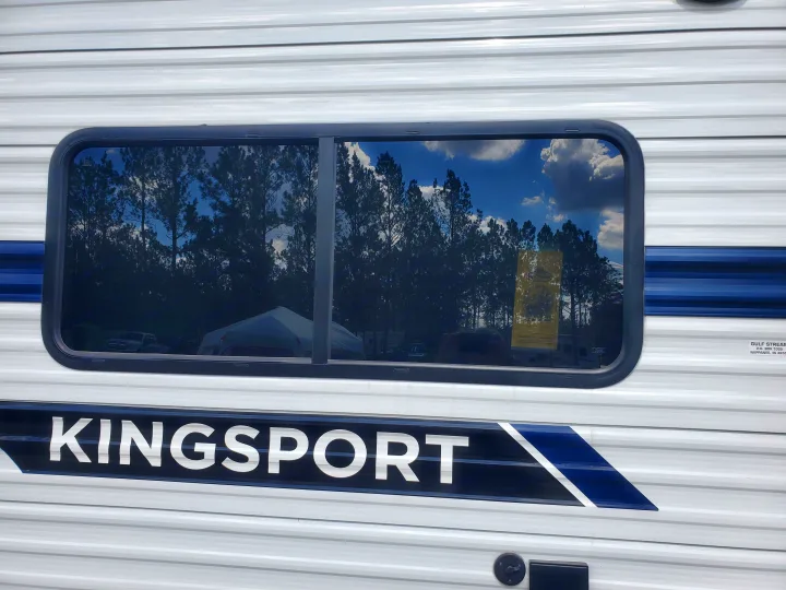 Kingsport RV rental side, white with blue stripes. Window reflects tall trees, blue sky, and clouds, perfect for nature getaways.. Gulf Stream Kingsport 2022