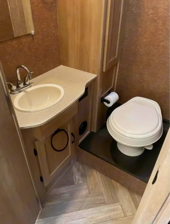 Inside an RV rental, this compact bathroom features a beige sink, light wood vanity, and a white toilet on an elevated platform.. Coachmen Freelander 2016