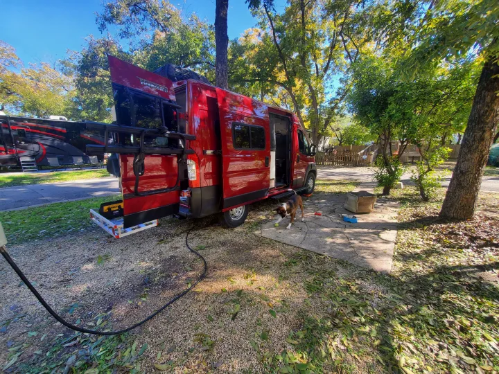 Red RV rental camper van parked at a sun-dappled campsite, with a playful dog on the ground.. Winnebago Solis Pocket 2022
