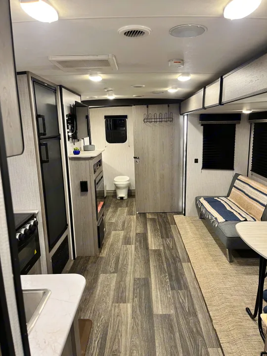 Modern RV rental interior showing a compact kitchen, living area with a futon, and a bathroom, featuring wood-look floors.. Heartland RVs Mallard 2021