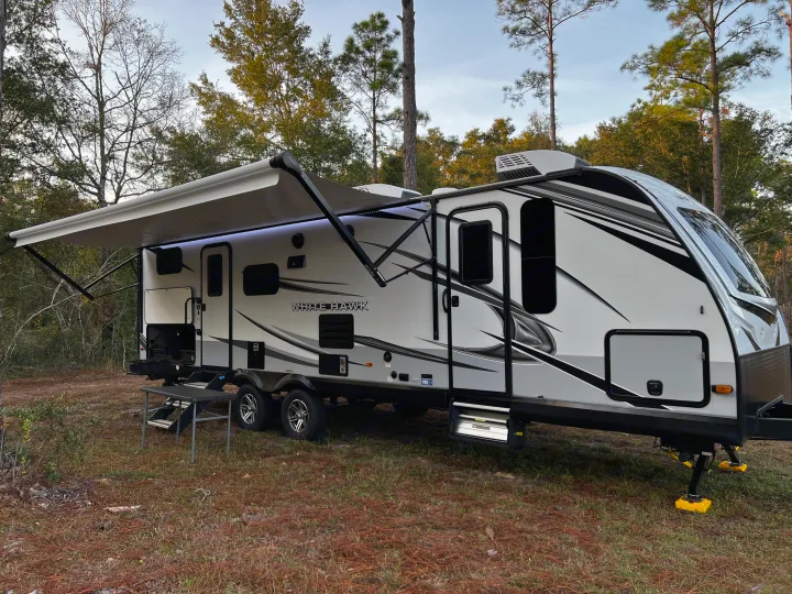 This sleek White Hawk RV rental, awning extended with lights, is parked amidst trees, perfect for a peaceful camping trip.. Jayco White Hawk 2022