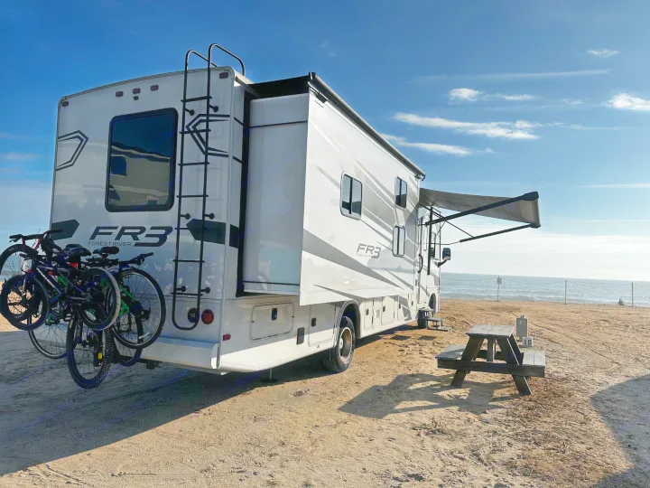 Enjoy an RV rental: a large motorhome with bikes & awning, set up on a sandy beach by the ocean with a picnic table.. Forest River Fr3 2021