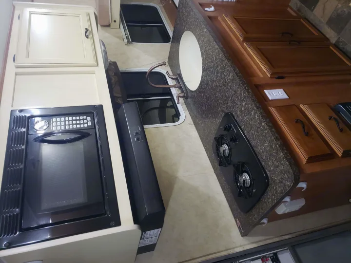 RV rental interior: a compact kitchen featuring a microwave, range hood, sink, two-burner stovetop, and wood cabinets.. Coachmen Apex 2012